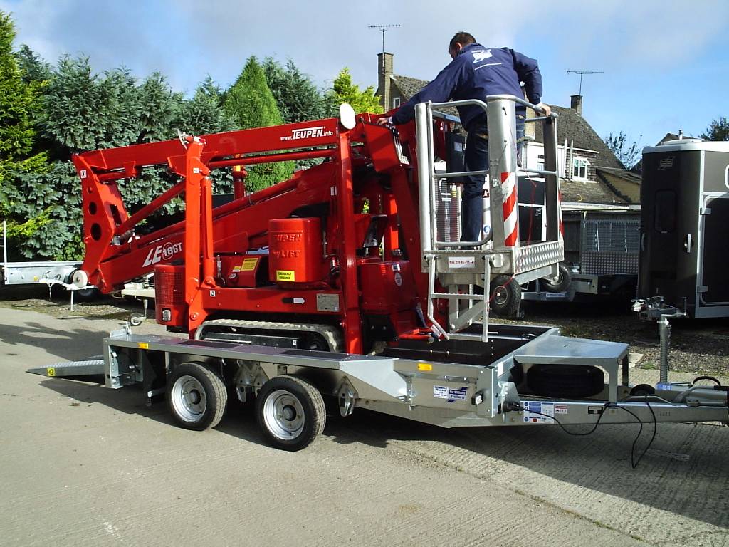 Red cherry picker on a trailer with a person on top, parked on a street.